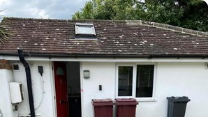 Une maison de plain-pied avec une porte rouge, deux poubelles et une petite fenêtre.