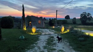 Une maison de campagne en pierre, avec un chemin de gravier menant à l’entrée, entourée de verdure et illuminée par de grandes lanternes.