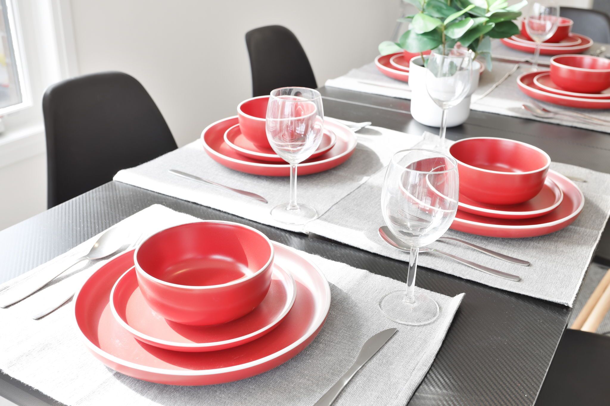 Una mesa de comedor dispuesta con platos, tazones y copas de vino rojos, sobre individuales grises.