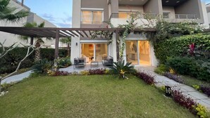 Une maison moderne à deux étages, dotée d’un patio couvert, d’un jardin bien entretenu et d’un intérieur visible, comprenant notamment un coin repas.