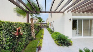 Un couloir extérieur moderne, agrémenté d’un chemin bordé d’une végétation luxuriante et de palmiers, qui mène à un bâtiment doté de grandes fenêtres et d’un espace abrité.