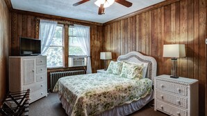Un dormitorio con paredes de madera, una cama con ropa de cama floral, dos lámparas de noche, un aparador, un televisor y un ventilador de techo.