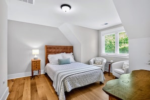 A bedroom with a sloped ceiling, a bed with a headboard, a bedside table with a lamp, a chair, a window with blinds, and a view of greenery outside.