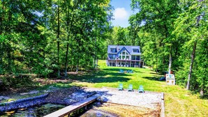 A large house with a deck, surrounded by trees and a dock extending into a clear water body.