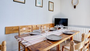 Un comedor con una mesa de madera dispuesta para cuatro, un televisor en la pared e impresiones botánicas enmarcadas.
