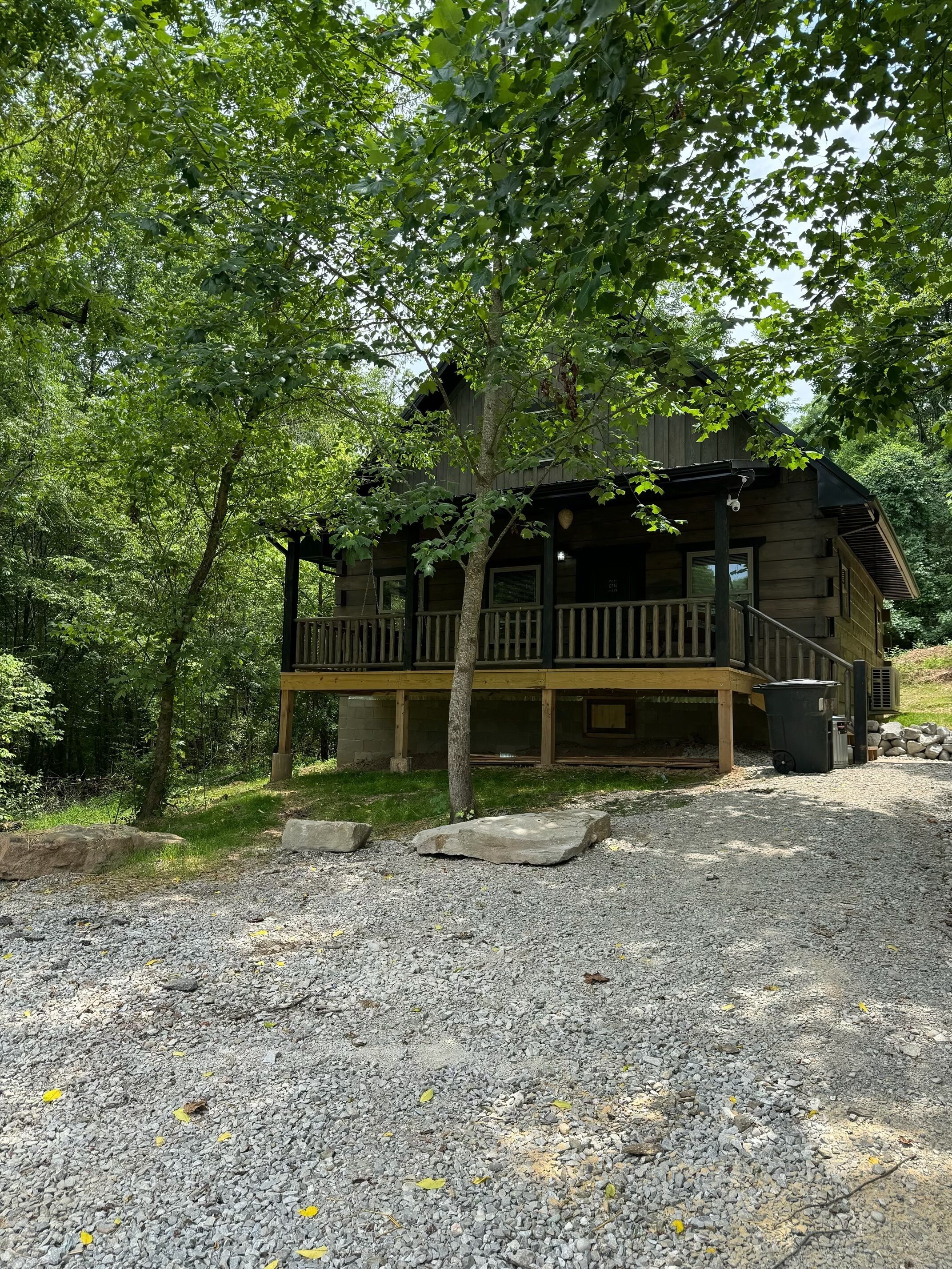 A wooden cabin with a porch, surrounded by trees and gravel ground.