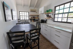 A compact kitchen with a round dining table, wooden chairs, a built-in oven, and a window with a view. - Potcake Villa — Stay with Purpose: Support Animal Welfare (cockburn town)