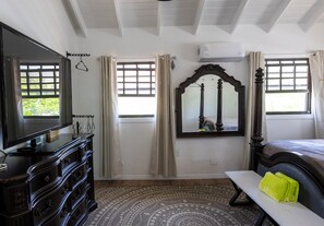 A bedroom with a bed, a dresser, a mirror, a television, and a window with curtains. - Potcake Villa — Stay with Purpose: Support Animal Welfare (cockburn town)