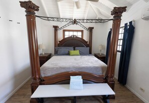 A four-poster bed with a wooden headboard and footboard, a white table in front, and two bedside tables with lamps. - Potcake Villa — Stay with Purpose: Support Animal Welfare (cockburn town)