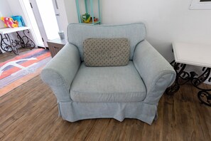 Un sillón gris claro con un cojín estampado en una habitación con suelo de madera y pared blanca.