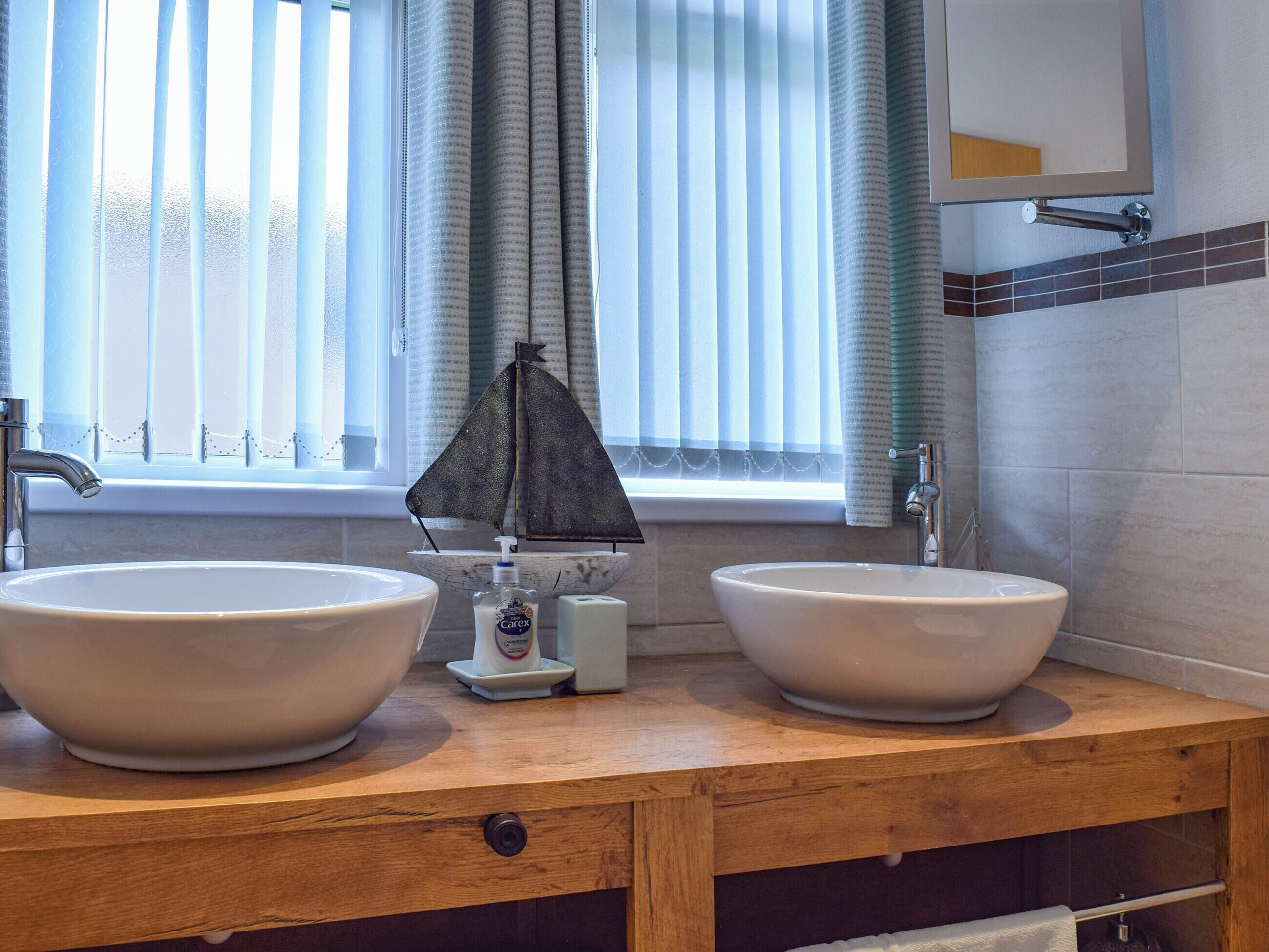 Une salle de bain avec deux grands lavabos blancs, un meuble-lavabo en bois, une petite maquette de bateau et une fenêtre avec des rideaux.
