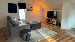 A cozy living room with a grey sofa, a round ottoman, a TV on a wooden stand, and a window with blinds. - Lovely house off Main Street (Hamilton)