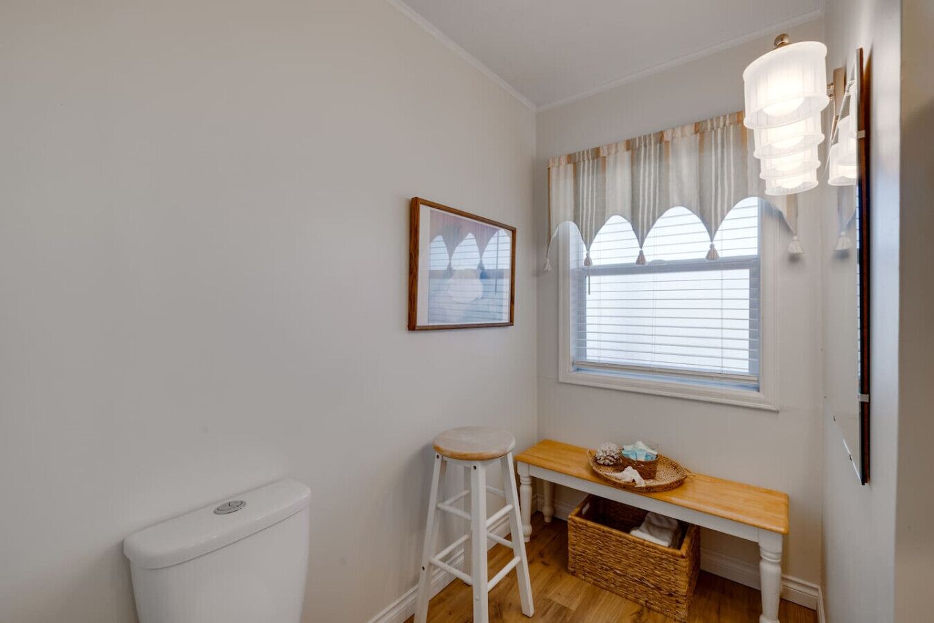 Un baño pequeño y limpio con inodoro, un taburete de madera, una cesta de mimbre y una ventana con cortinas.