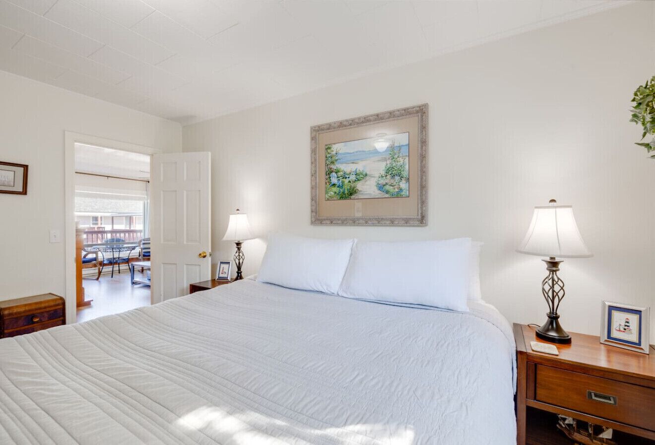 Une chambre avec un grand lit, deux tables de chevet avec des lampes, un tableau encadré au mur et une porte donnant sur une autre pièce.