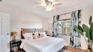 Une chambre à coucher comprenant un lit, une table de chevet, un ventilateur de plafond, une fenêtre avec des rideaux et une plante en pot.