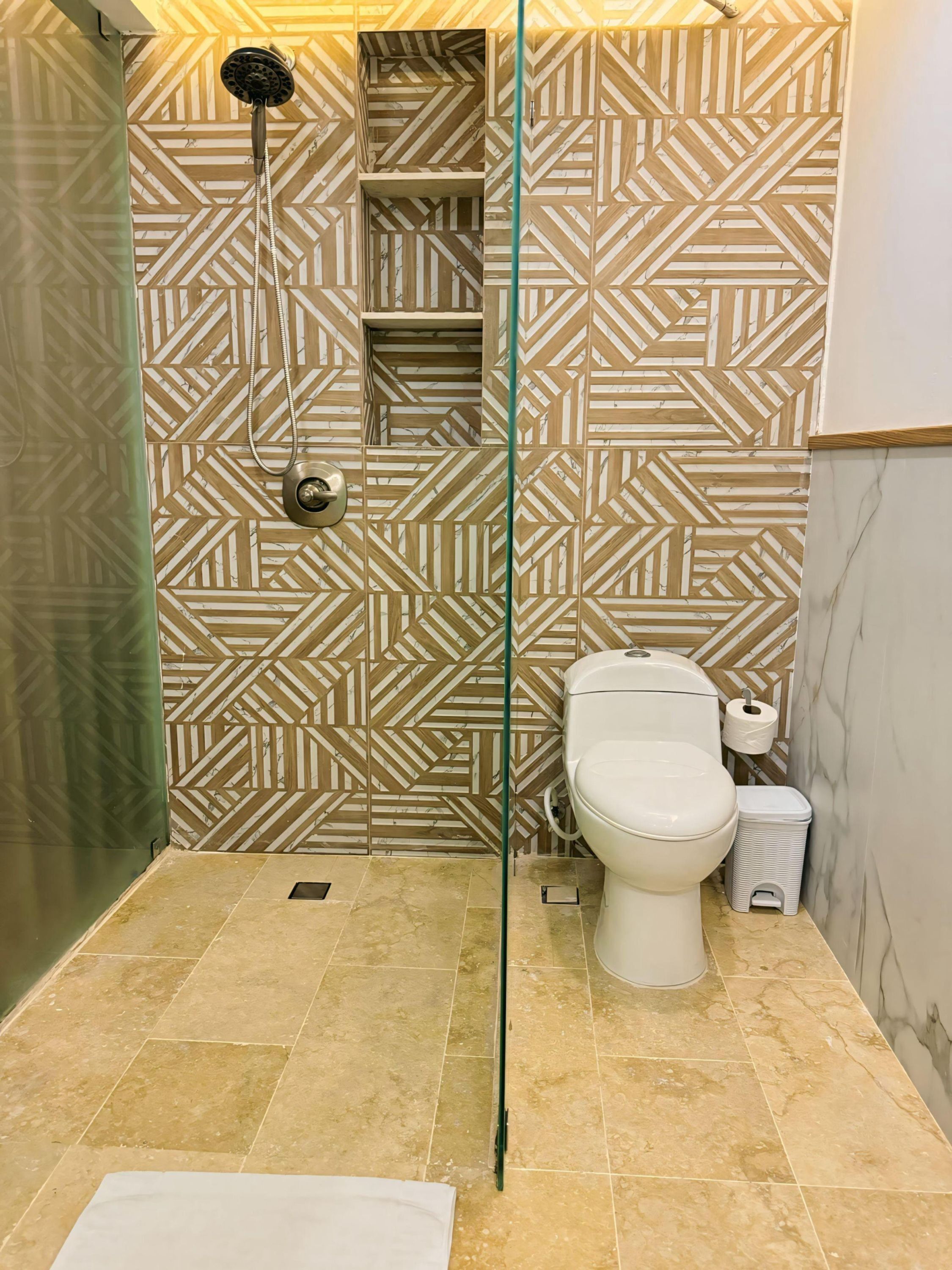 A bathroom with a geometric patterned wall, a glass shower enclosure, a toilet, and a trash bin.