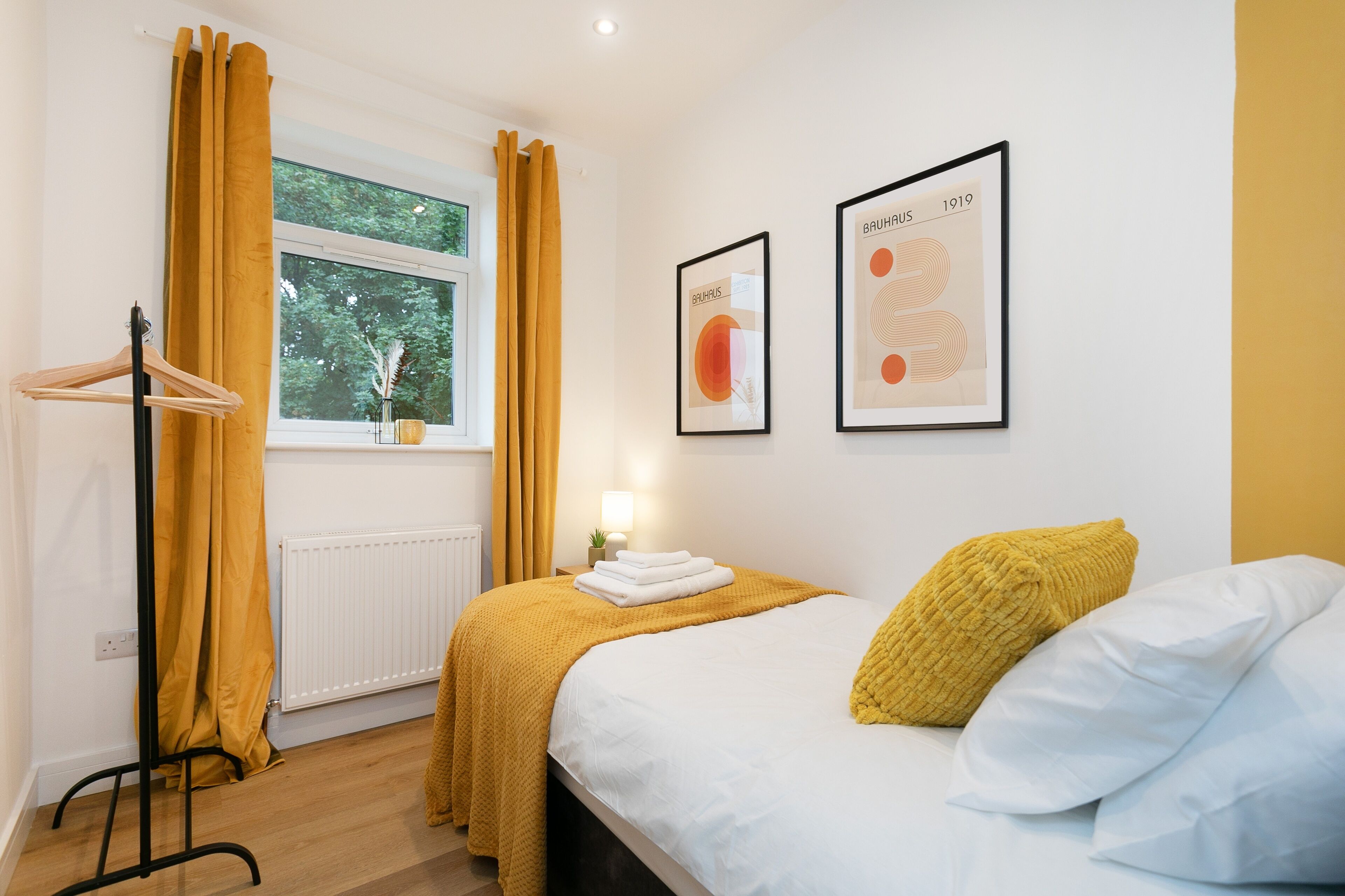 A bedroom with a bed, a nightstand, a window with curtains, and two framed artworks on the wall.