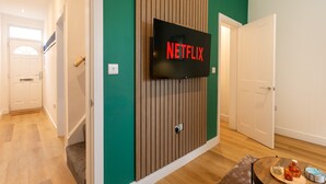 A modern living room with a mounted TV displaying the Netflix logo, a wooden panel wall, and a glass coffee table with items on it.