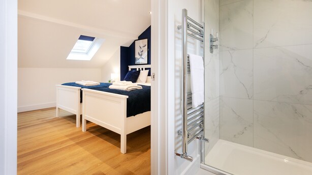 A bedroom with a bed, a bedside table, a nightstand, a window, and a shower area with a glass door.