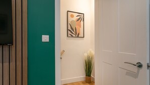 A hallway with a wooden floor, a green wall, a framed abstract art piece, a potted plant, and a door with a handle.