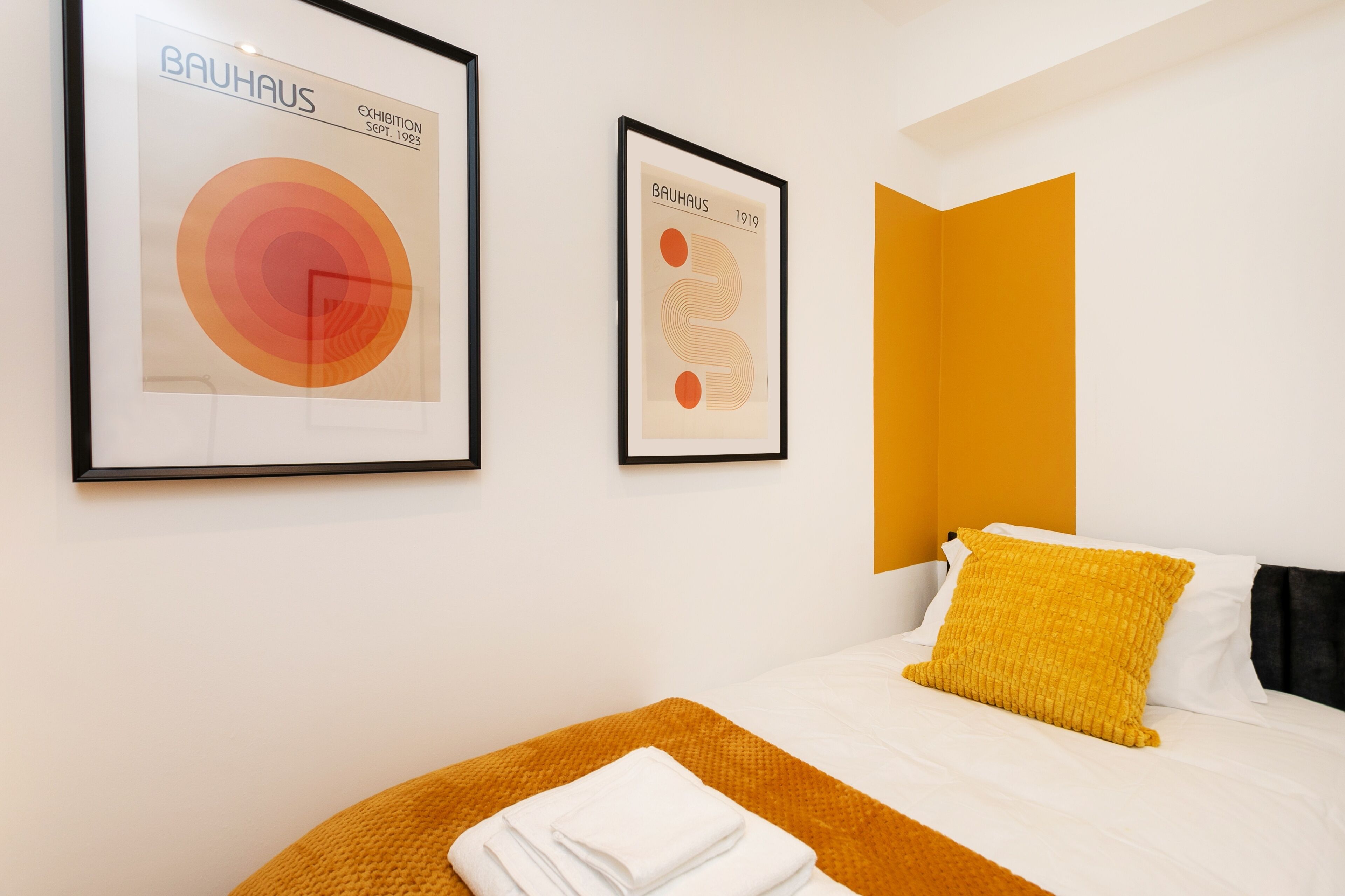 A hotel room with a bed, two framed artworks on the wall, and a bright orange accent wall.