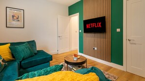 A modern living room with a green accent wall, a flat-screen TV displaying 'NETFLIX', a green sofa with yellow cushions, and a wooden panel wall.