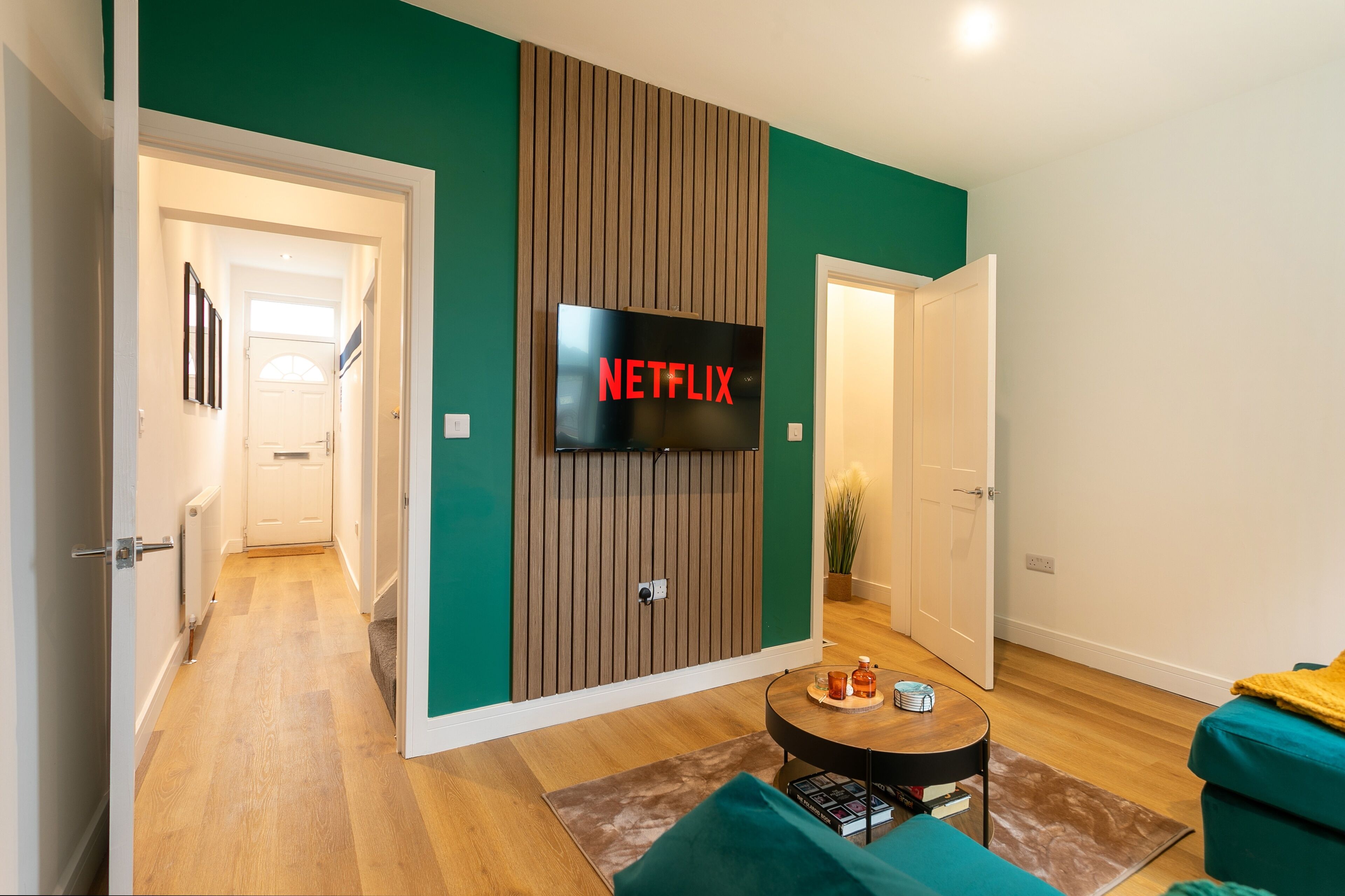 A modern living room with a flat-screen TV mounted on a wooden panel wall, a teal sofa, a coffee table, and a hallway leading to another room.