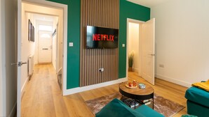 A modern living room with a flat-screen TV mounted on a wooden panel wall, a teal sofa, a coffee table, and a hallway leading to another room.