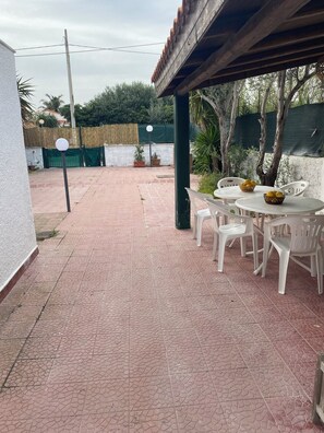 Un patio couvert avec un sol carrelé, des chaises et des tables en plastique blanc. On aperçoit, à l’arrière-plan, des plantes en pot et une clôture verte.