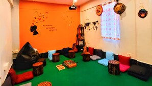 Una habitación con alfombra verde, paredes naranjas y un mural con la silueta de una persona. Hay taburetes de madera, cojines y una guitarra.