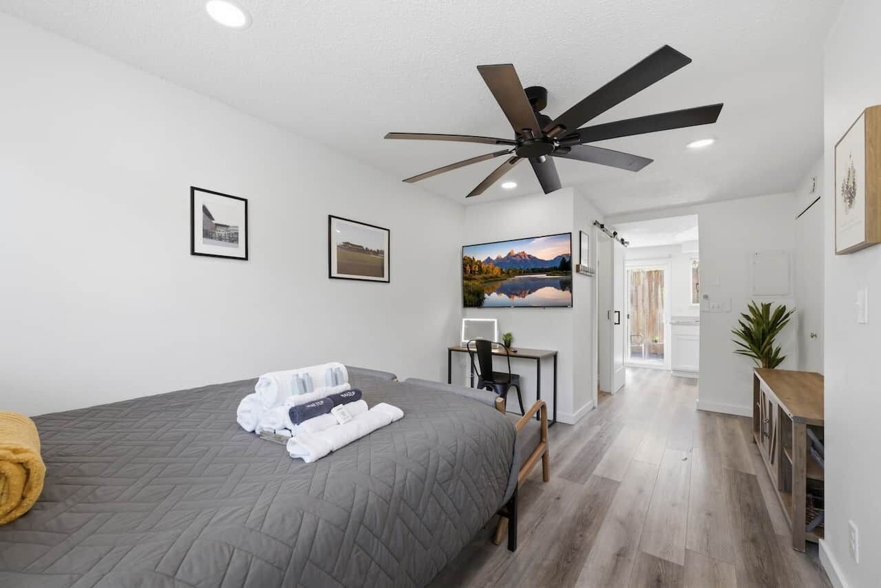 A hotel room with a bed, a desk, a chair, a ceiling fan, and a view of a mountain landscape.
