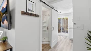 A modern interior with a mounted TV, a wooden shelf with a framed picture, a glass shower enclosure, and a view through an open door to a balcony.