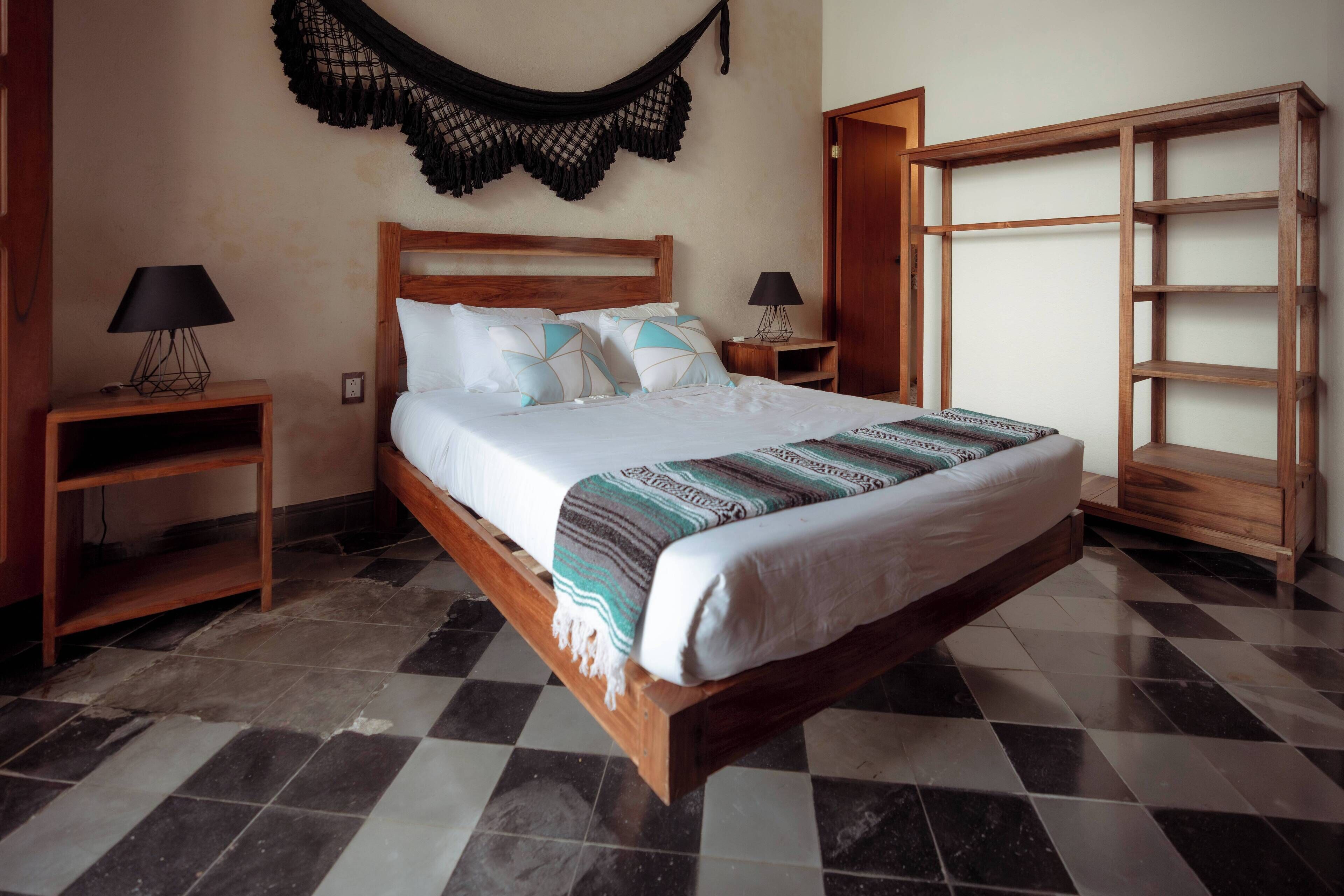 A neatly made bed with a wooden headboard, two bedside tables with lamps, a hanging macrame wall decor, and a wooden shelving unit.