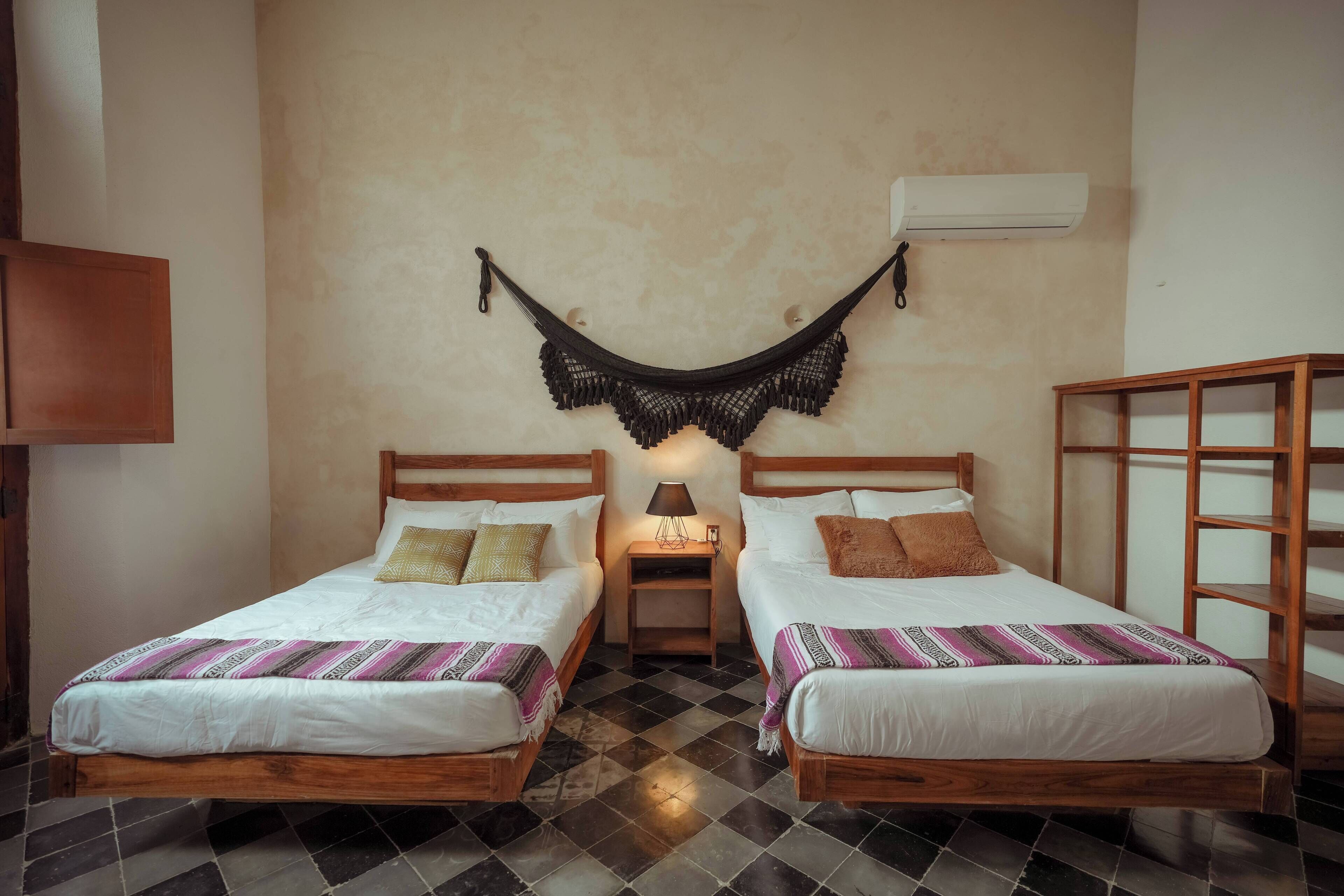 Una habitación con dos camas de madera, una hamaca, una mesita con una lámpara y un armario de madera.