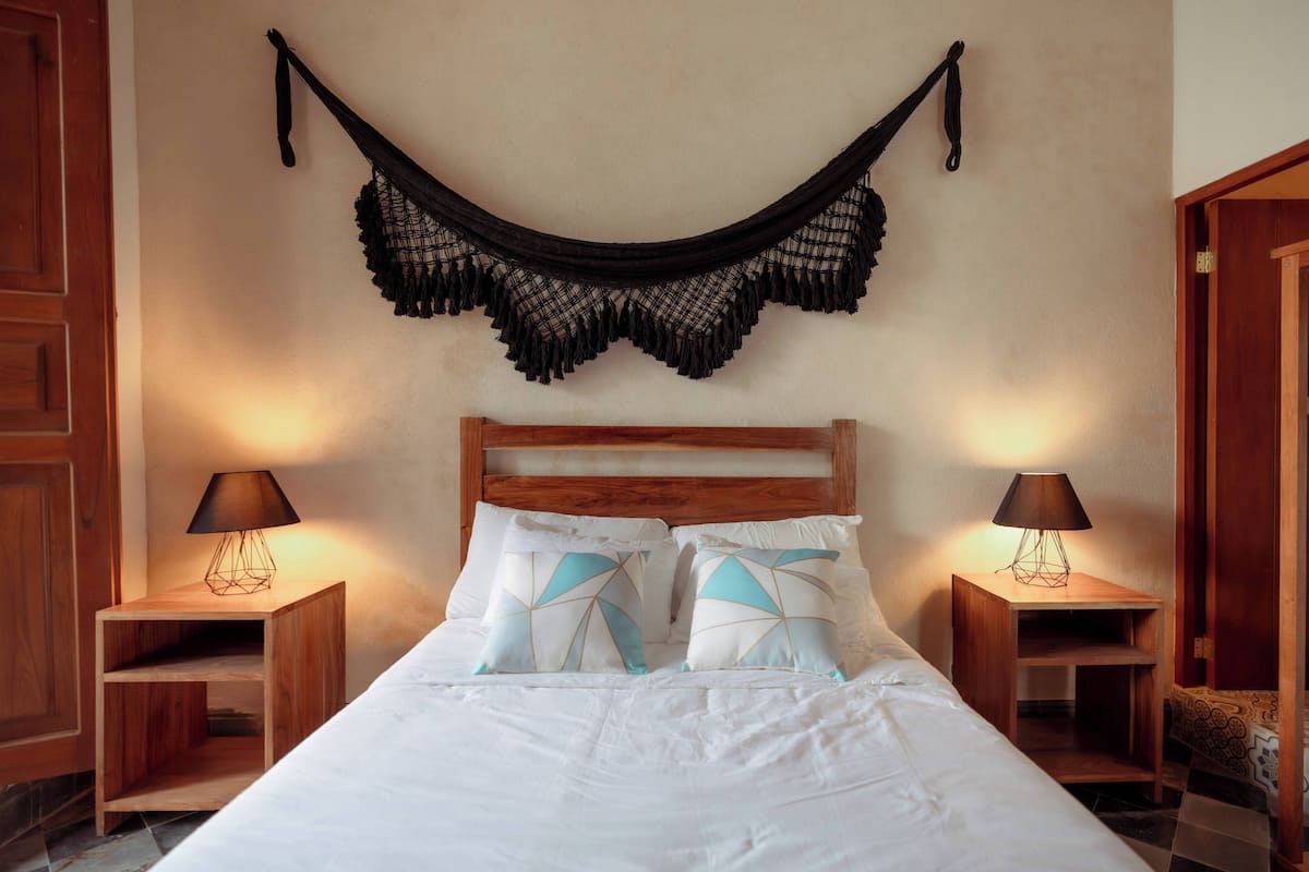 Una cama bien hecha con sábanas blancas y almohadas de estampado geométrico, flanqueada por mesitas de noche de madera con lámparas, en una habitación con cabecera de madera y una hamaca negra colgando.