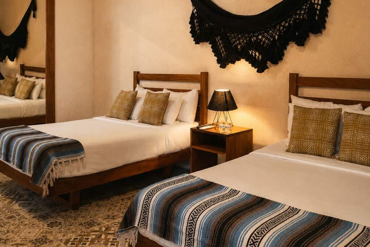 Habitación de hotel con dos camas, cabecero de madera, alfombra estampada, un textil negro colgante y una mesita de noche con lámpara.