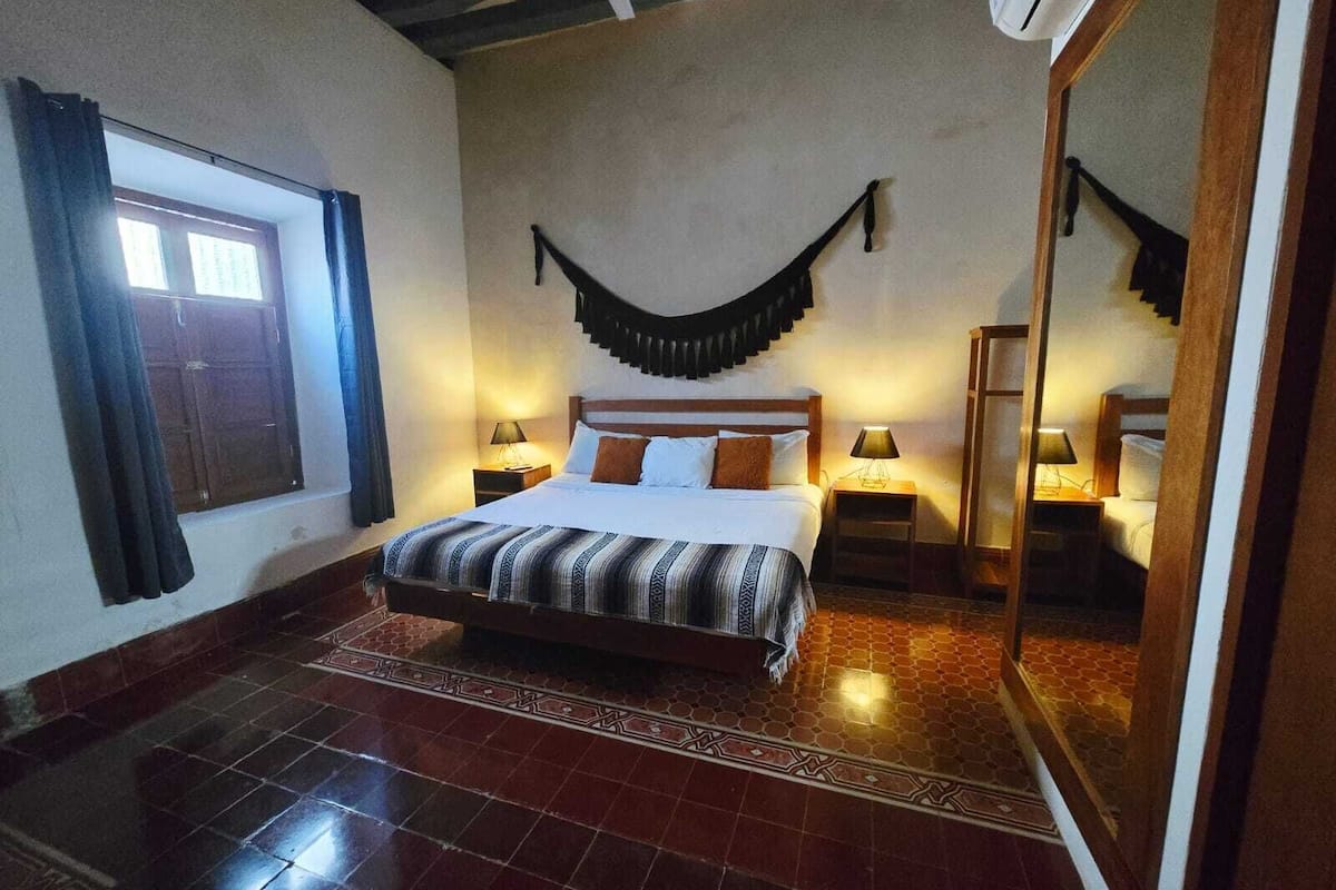 Una habitación de hotel con una cama grande, dos mesitas de noche con lámparas, un ventanal con cortinas y un suelo de baldosas.
