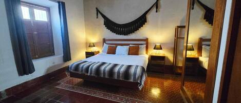 A hotel room with a large bed, two bedside tables with lamps, a window with curtains, and a tiled floor.