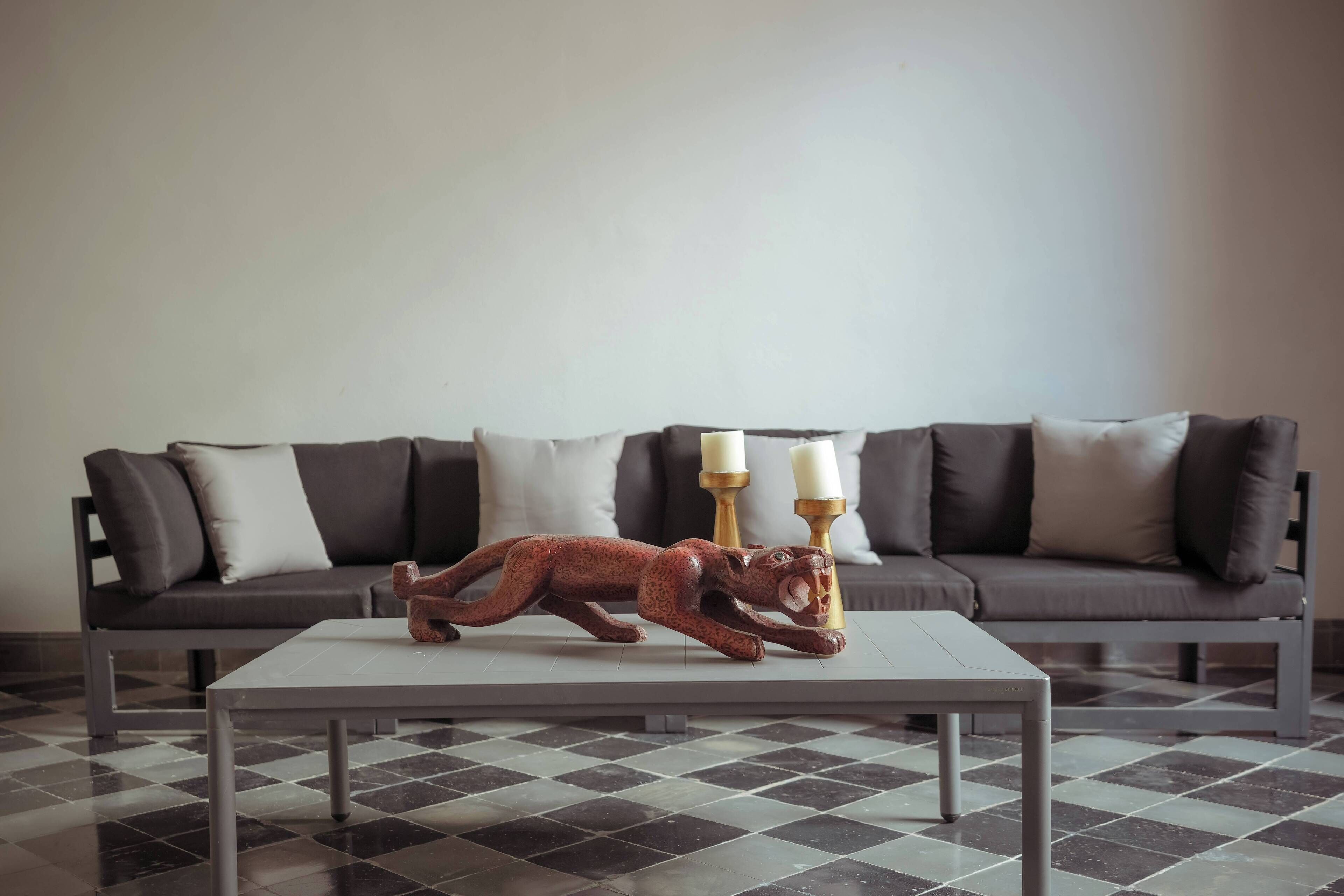 Ein modernes Wohnzimmer mit Schachbrettmuster-Boden, einer Couch mit Kissen und einer dekorativen Holzfigur eines Tieres auf einem Couchtisch.