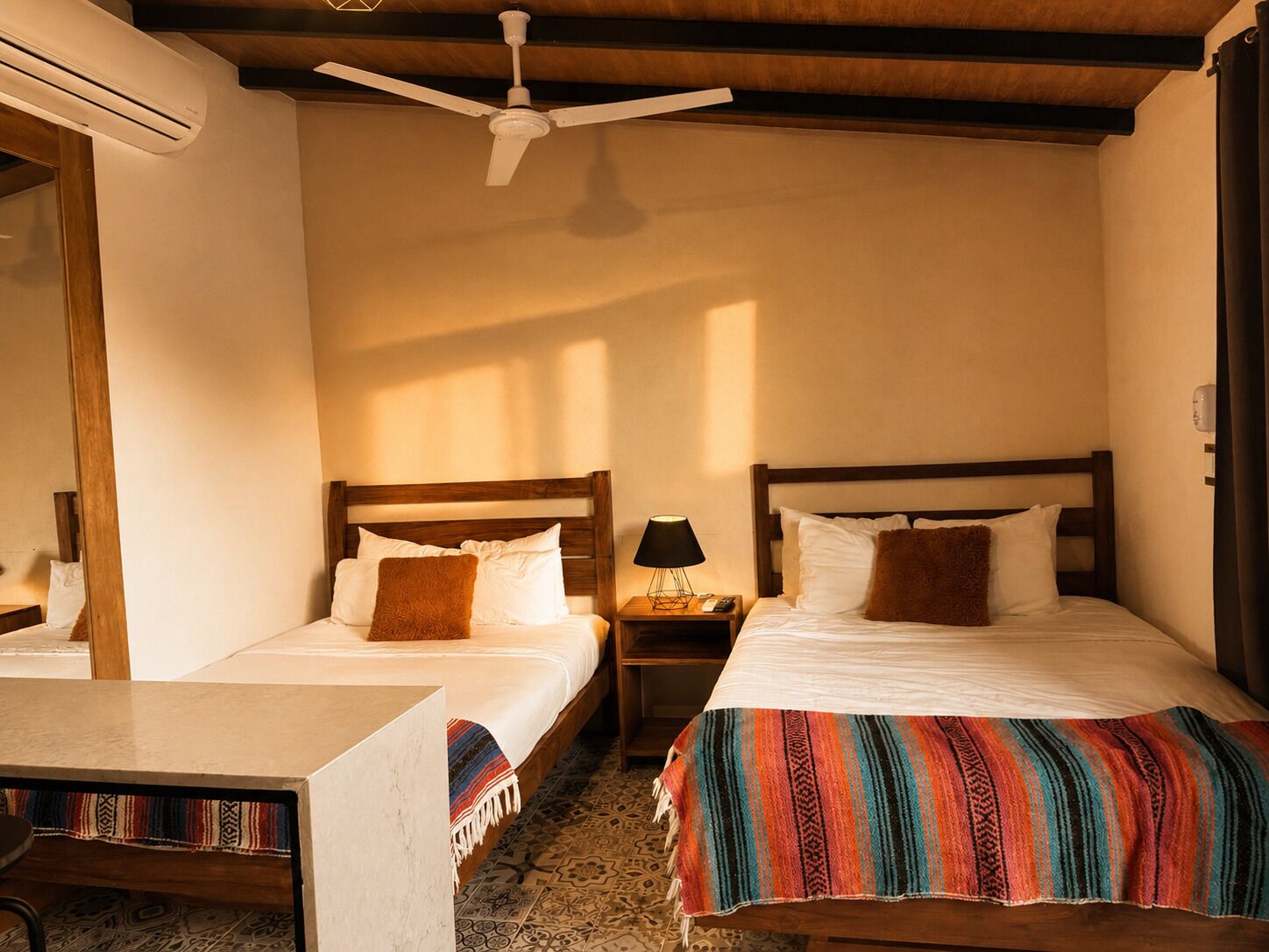 Habitación de hotel con dos camas, un escritorio, una mesita de noche, un ventilador de techo y una ventana.