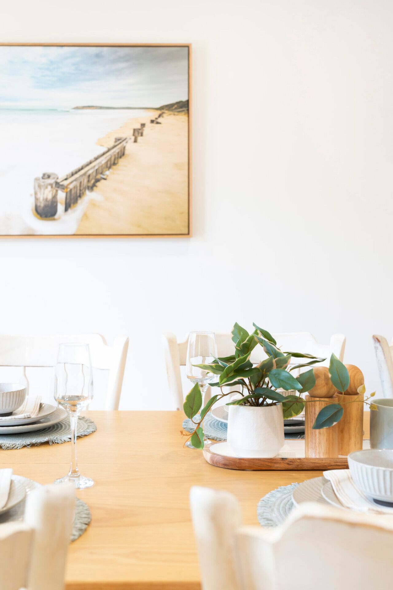 A dining table set with white plates, bowls, and glasses, a potted plant, and a wooden tray.