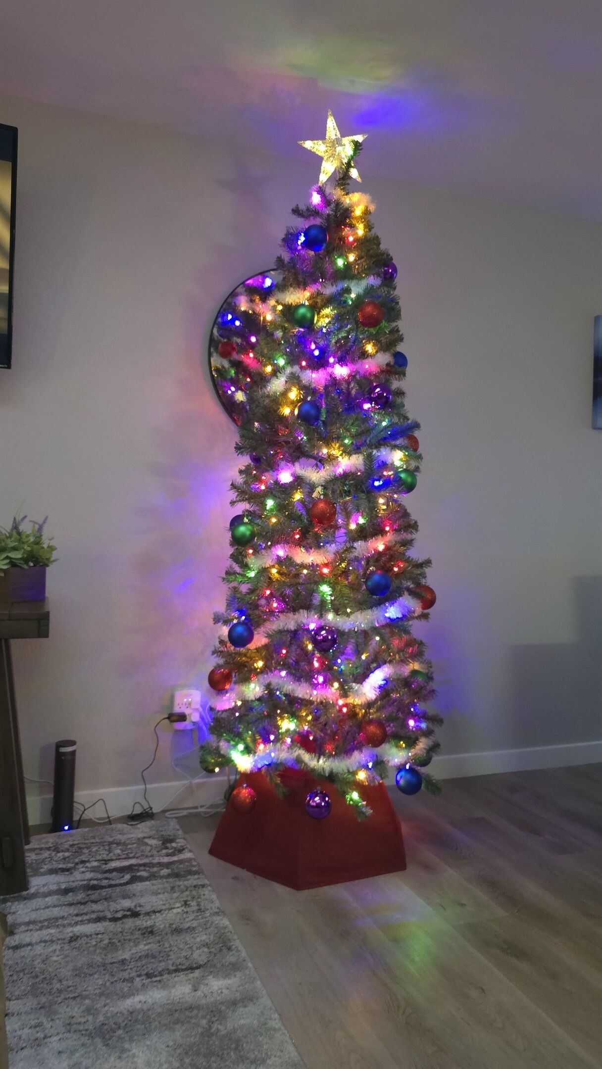 Een kerstboom met kleurrijke lichtjes en versieringen, geplaatst in een kamer met een houten vloer en een klein rode voetstuk.