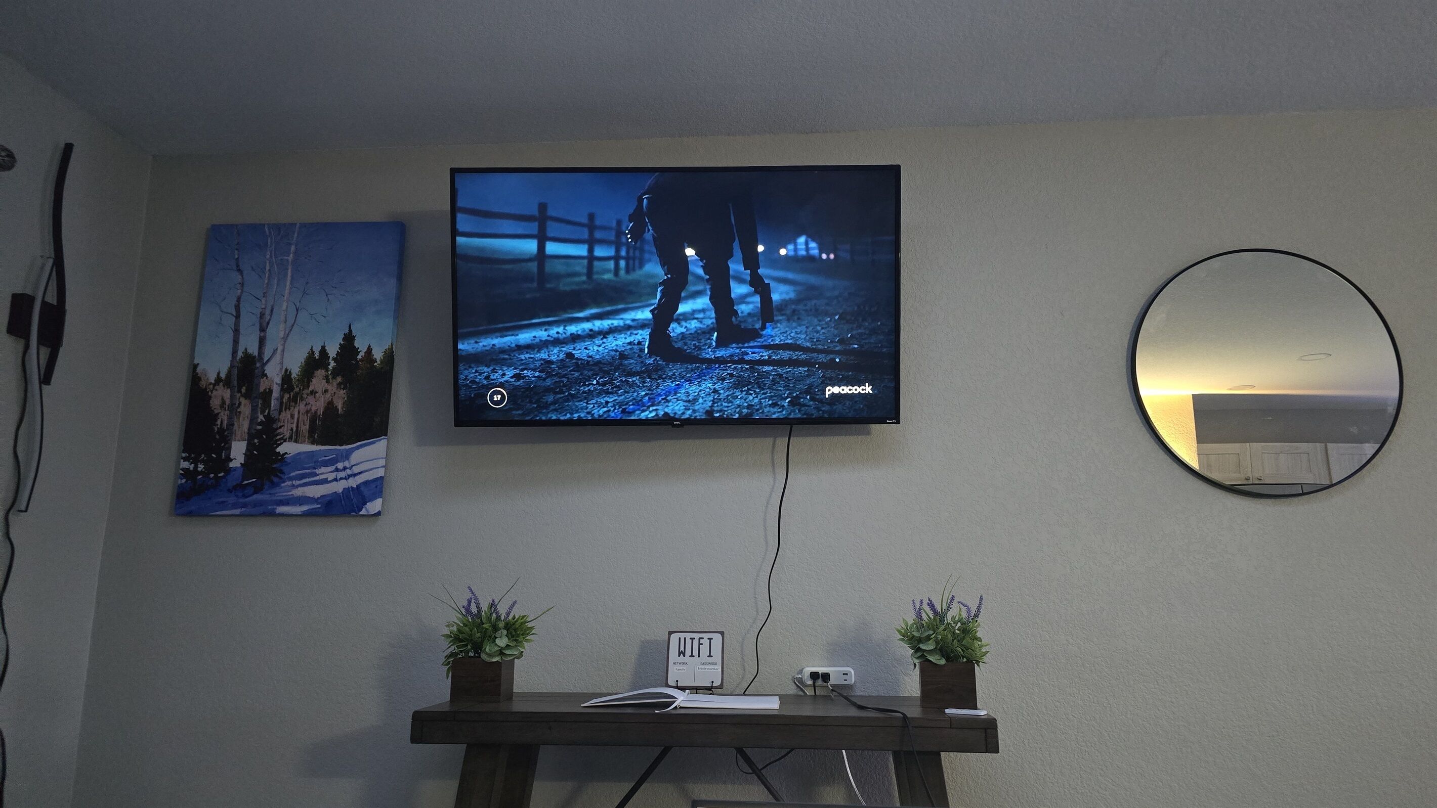 Ein an der Wand befestigter Fernseher zeigt eine Szene mit zwei Personen, ein Gemälde von verschneiter Landschaft, ein runder Spiegel und ein kleiner Tisch mit Pflanzen und einem Schild, das kostenloses WLAN anzeigt.