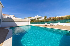 Une piscine dotée d’une terrasse en bois et de chaises longues, entourée de bâtiments blancs et surplombée par un ciel bleu azur.