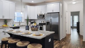 A modern kitchen with white cabinets, a stainless steel refrigerator, and a central island with stools. - Surfside Clubhouse: Steps to Beach & Heated Pool and Arcade Games! (Surfside Beach)