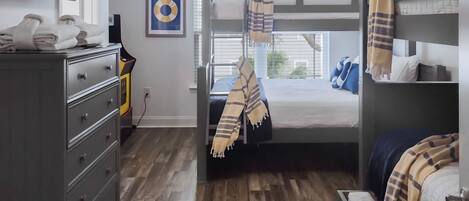 Una habitación con una cama litera, un escritorio con cajones, un televisor en la pared y una ventana con persianas.