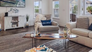 A living room with a large TV, a sofa, a coffee table, and a view of the beach. - Surfside Clubhouse: Steps to Beach & Heated Pool and Arcade Games! (Surfside Beach)