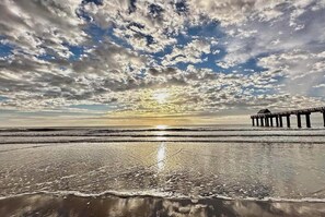 A beach with a pier extending into the water, waves gently lapping at the shore, and a partly cloudy sky above. - Surfside Clubhouse: Steps to Beach & Heated Pool and Arcade Games! (Surfside Beach)