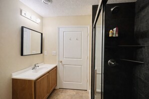 Un baño con lavamanos blanco, armarios de madera, un espejo y una ducha con azulejos oscuros.