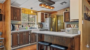 A kitchen with wooden cabinets, a large island with stools, a refrigerator, and a sign that reads "SUNSHINE". - Hardscrabble Mesa | Pine Trees | Wildlife | ADA (Pine)